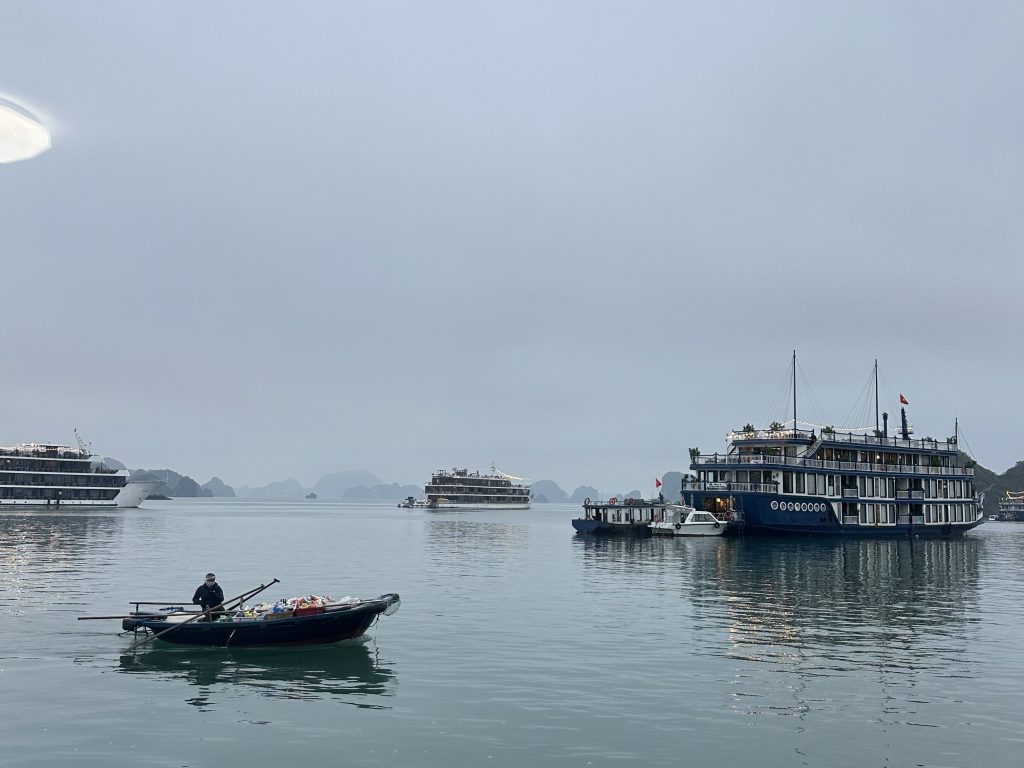 Breathtaking view of foggy Ha Long Bay showcasing several cruises anchored amidst towering limestone islands, with a passing boat vendor offering local goods.