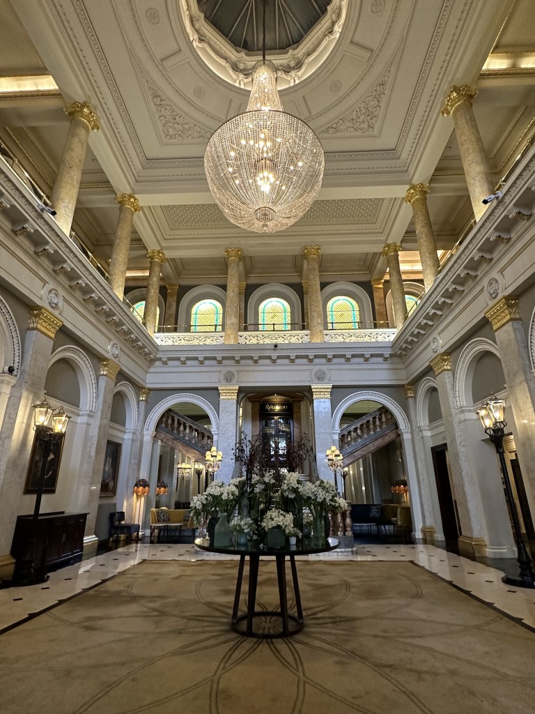 Interior view of a grand hall featuring high ceilings, ornate chandeliers, and decorative pillars. A circular table with floral arrangements is positioned in the center, surrounded by elegant furnishings and large windows.