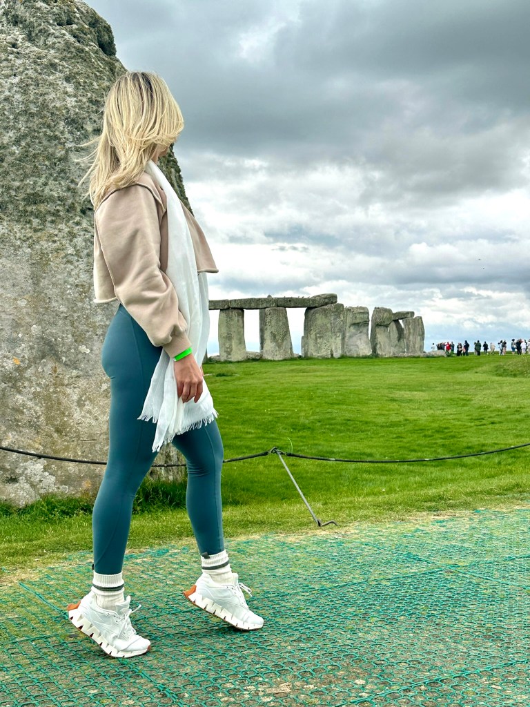 A person standing near Stonehenge, wearing a light jacket and leggings, with long blonde hair blowing in the wind, against a cloudy sky.