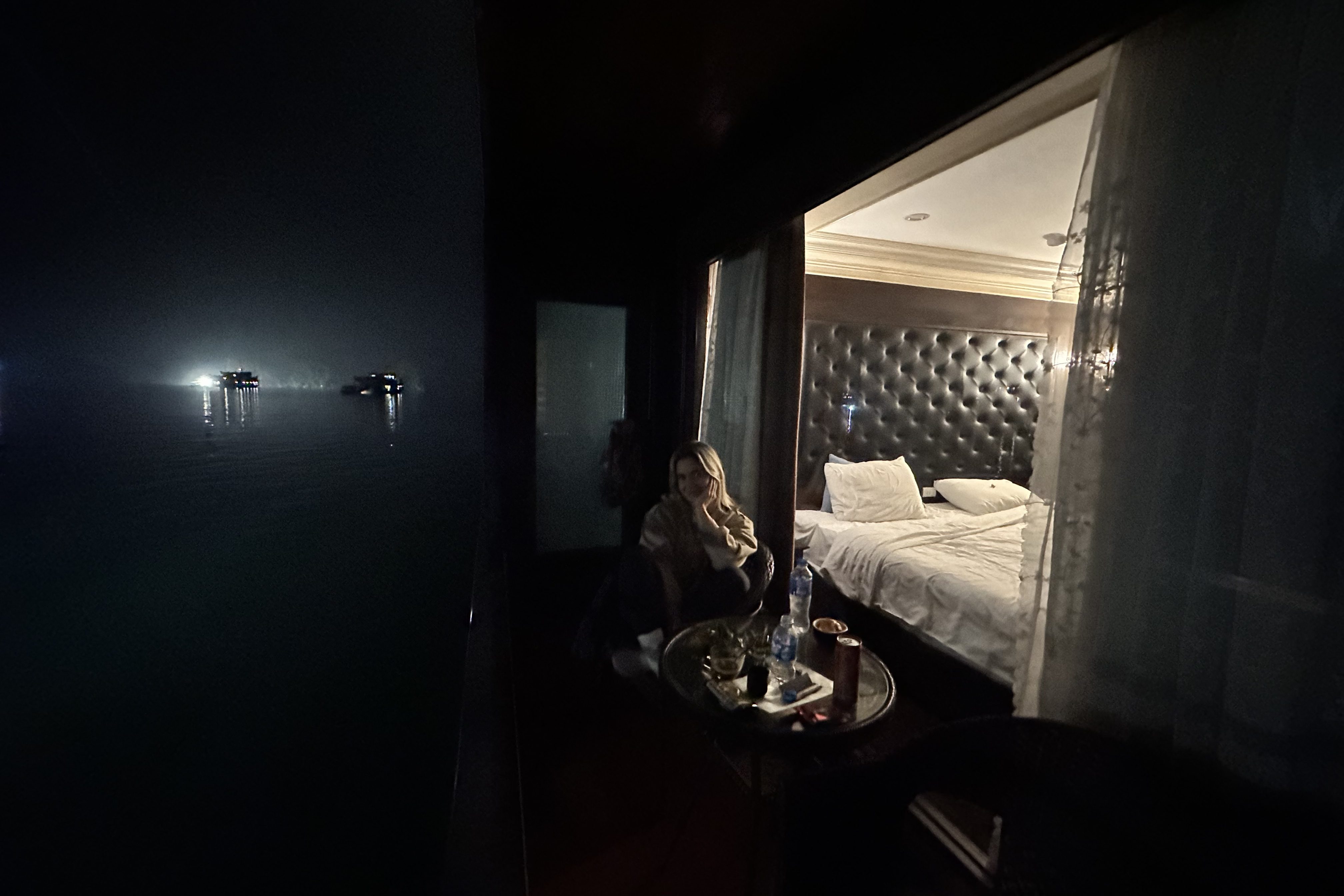 Nathalie sitting on the chairs of the balcony at night on the ship in Ha Long Bay, relaxing while overlooking the calm waters and the silhouette of the limestone islands under the night sky.
