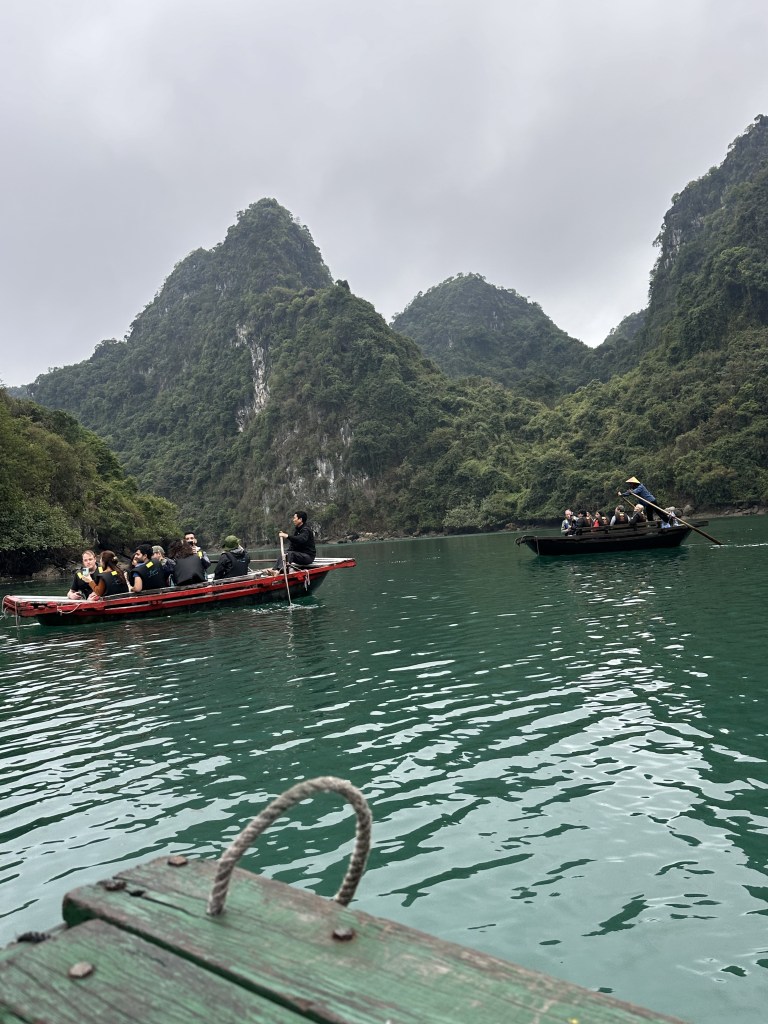Traditional row boats gliding through the tranquil waters of Ha Long Bay, transporting passengers to the entrance of the Dark & Bright Cave, surrounded by stunning limestone formations.