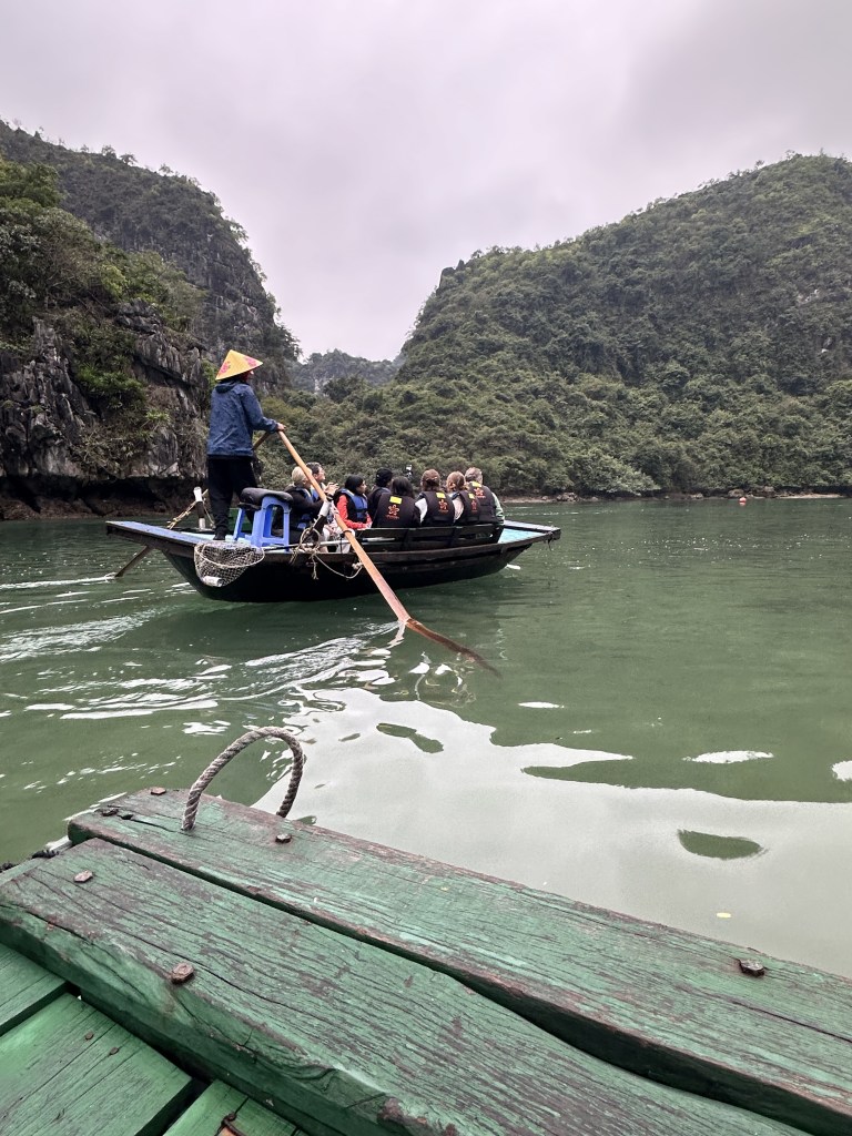 Traditional row boats gliding through the tranquil waters of Ha Long Bay, transporting passengers to the entrance of the Dark & Bright Cave, surrounded by stunning limestone formations.