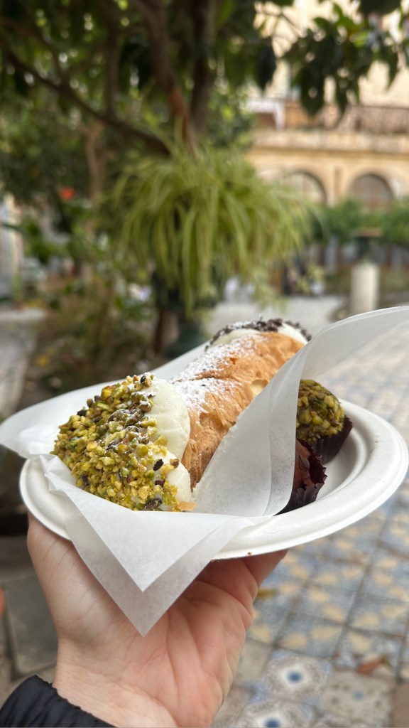 A hand holding a plate with two desserts, including a pistachio-coated pastry and a chocolate muffin, set against a background of greenery.