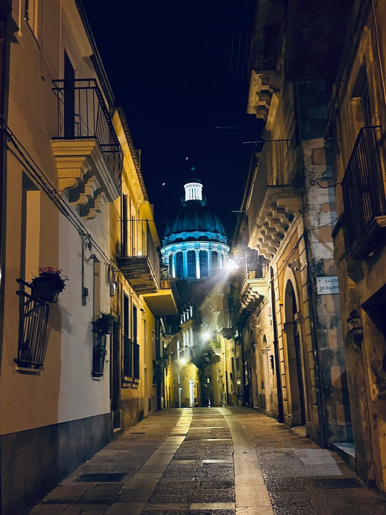 Narrow alleyway at night with illuminated buildings and a prominent dome in the background.