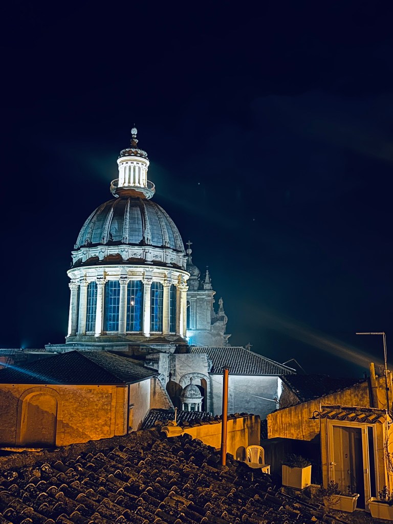 Illuminated dome of a historic building at night, viewed from rooftop, with dark sky in the background.