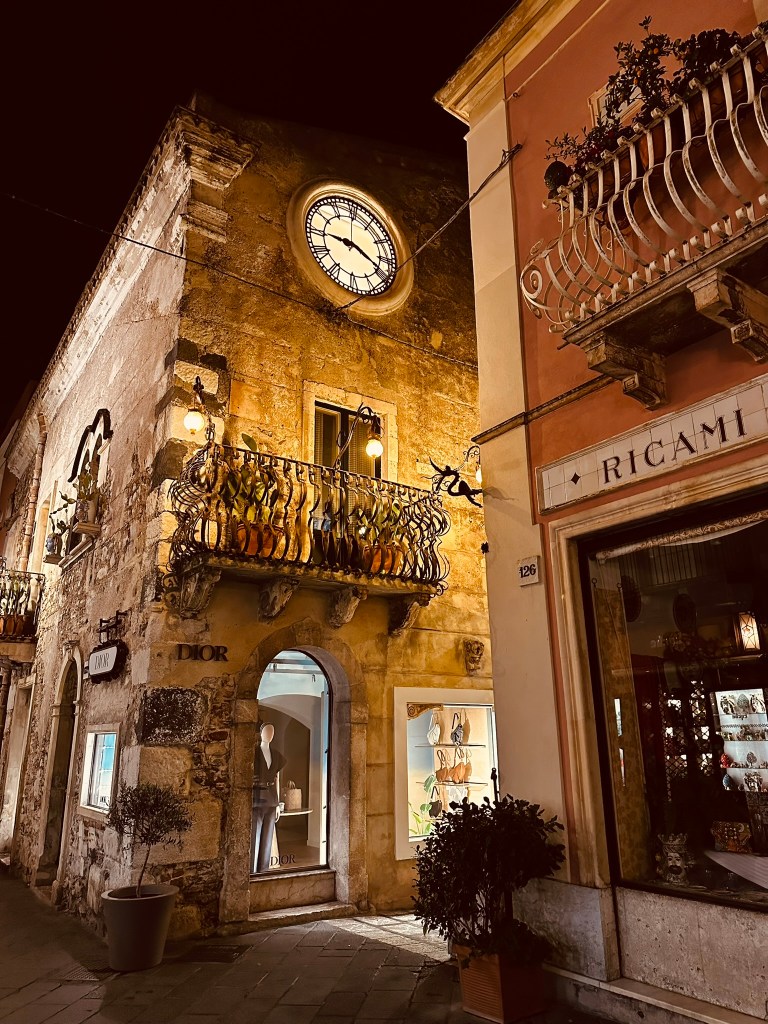 Illuminated streetscape at night featuring a vintage clock on a stone building, a boutique storefront with Dior branding, and ornamental balconies adorned with plants.