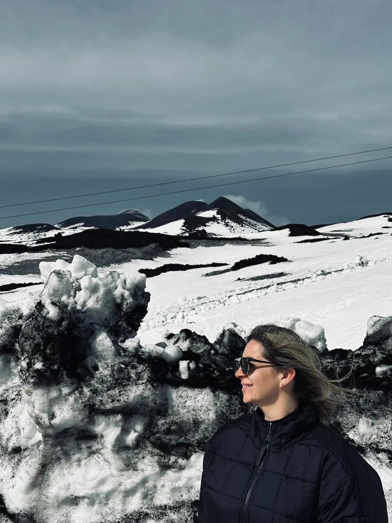 A woman in a black jacket stands in front of snow-covered volcanic mountains under a gray sky. She is wearing sunglasses and appears to be enjoying the scenic view.