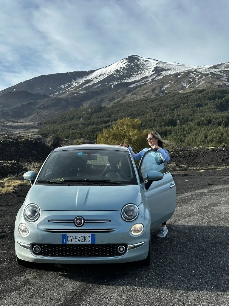 A light blue Fiat 500 parked on a gravel road with a woman standing next to it against a backdrop of a snow-capped mountain.