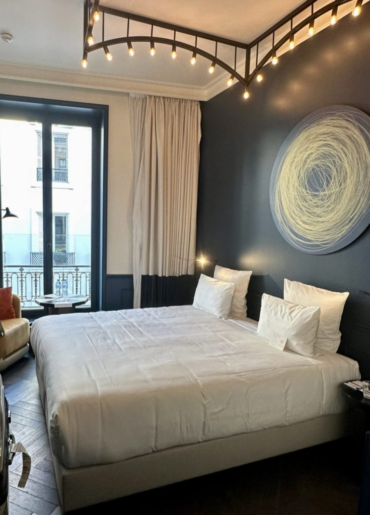 A modern bedroom featuring a large white bed with fluffy pillows, a stylish light fixture above, and large windows allowing natural light to enter. Neutral-colored curtains frame the window, and a cozy seating area with a colorful chair is visible in the corner.