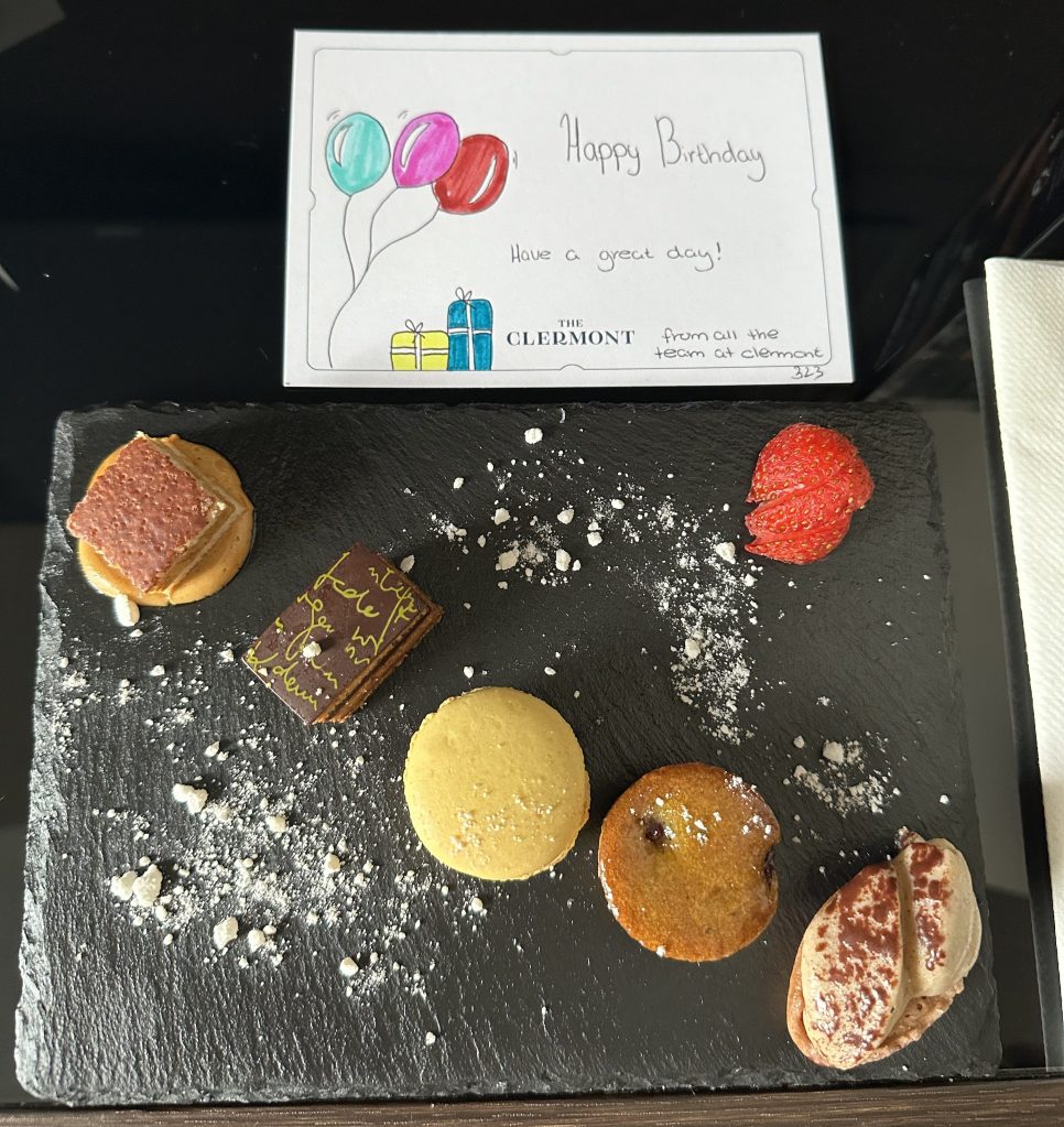 A black slate platter displaying an assortment of small desserts, including a macaron, a chocolate treat with writing, a yellow cookie, and a strawberry. A colorful birthday card with balloons and a gift illustration rests on the side.