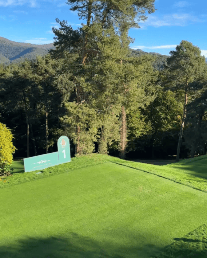 A scenic view of the first tee on a golf course, featuring a vibrant green grass area and a sign indicating tee number one, surrounded by tall trees and mountains in the background.