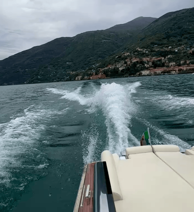 A view from a boat showing water splashing behind while cruising on a lake surrounded by mountains and a waterfront village in the background under a cloudy sky.