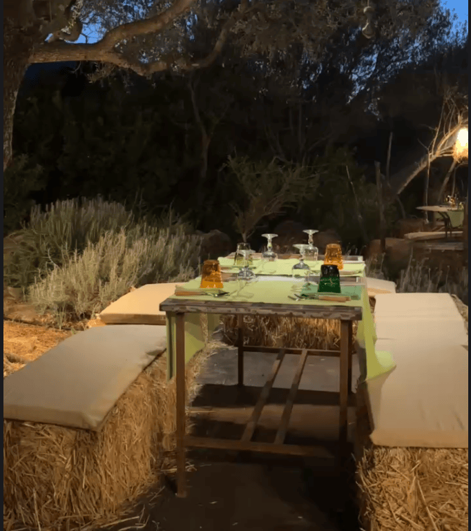 Outdoor dining setup with a wooden table covered in a green tablecloth, featuring glassware and colorful jelly cups, surrounded by straw seating and lush greenery.