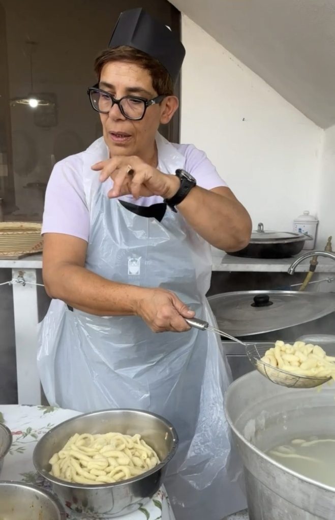 A woman wearing a white apron and glasses is holding a slotted spoon with pasta above a large pot, while standing in a kitchen setting.