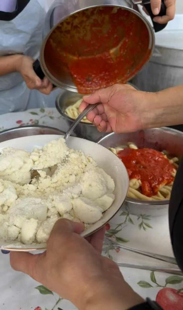 A person holding a bowl of ricotta cheese with a spoon, while another hand pours tomato sauce from a pot into a bowl of pasta in the background.
