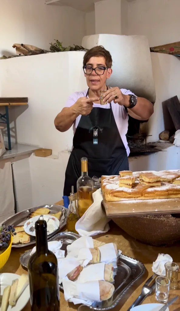 A woman in an apron holding a small glass and gesturing, surrounded by a table filled with various dishes and bottles in a rustic kitchen setting.