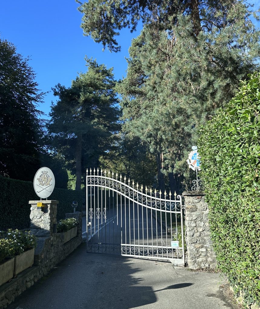 A decorative wrought-iron gate surrounded by tall green trees, leading to a driveway behind stone walls and hedges.