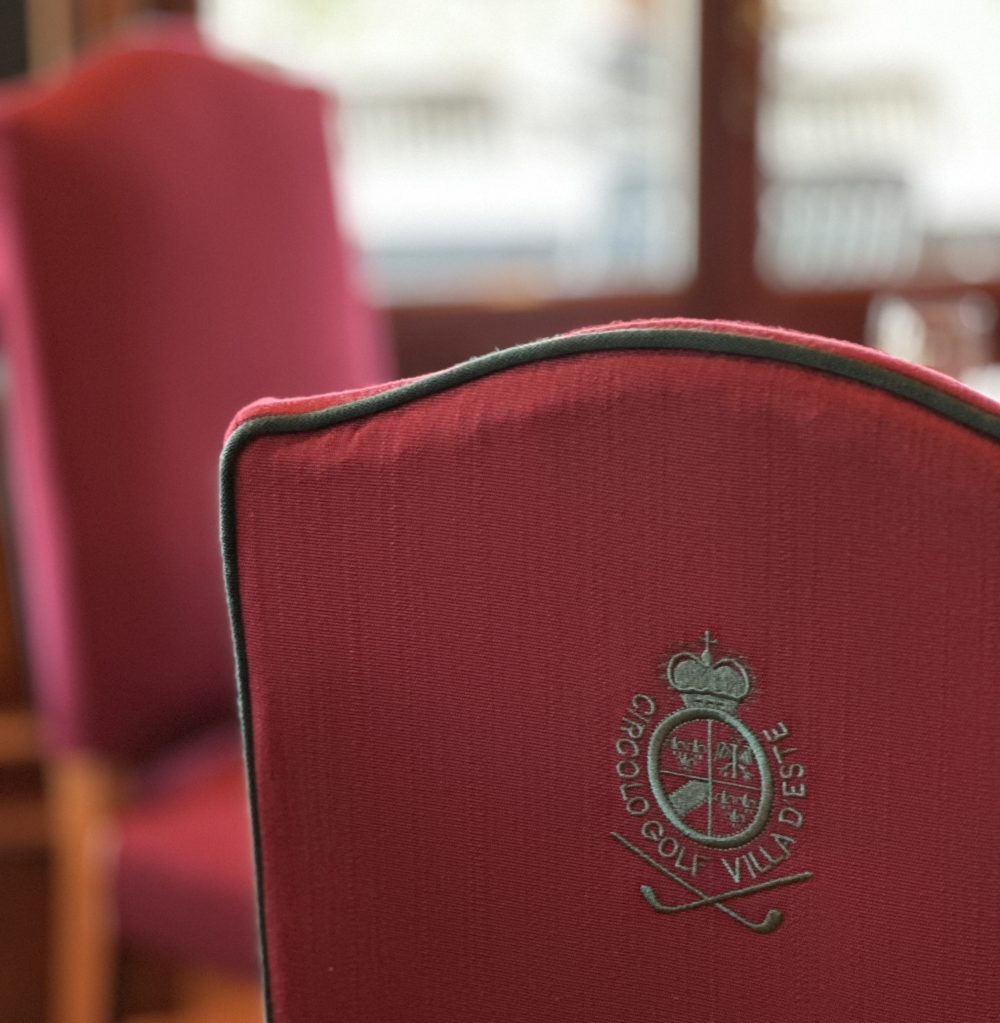Close-up of a red upholstered chair back featuring an embroidered logo with text 'Circolo Golf Villa d'Este' and golf clubs.