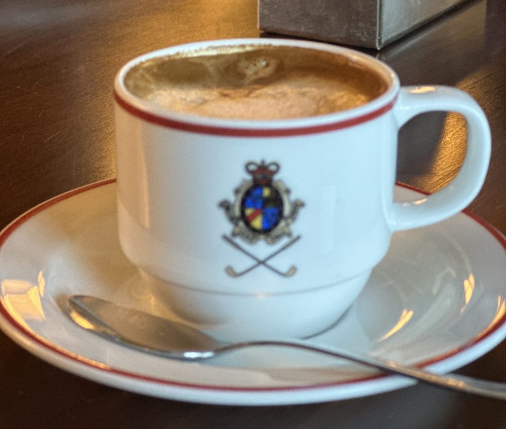 A close-up of a decorated coffee cup with a brass emblem, sitting on a small saucer, alongside a silver spoon.