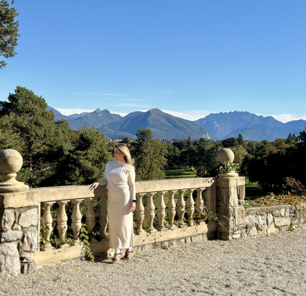 A woman in a light-colored dress stands by a stone railing overlooking a picturesque landscape with mountains and trees under a clear blue sky.