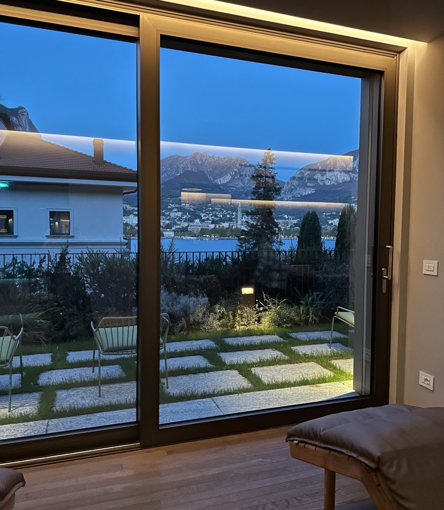 A modern window view showcasing a tranquil garden with stone pathways, greenery, and chairs, overlooking a lake and mountains during dusk.
