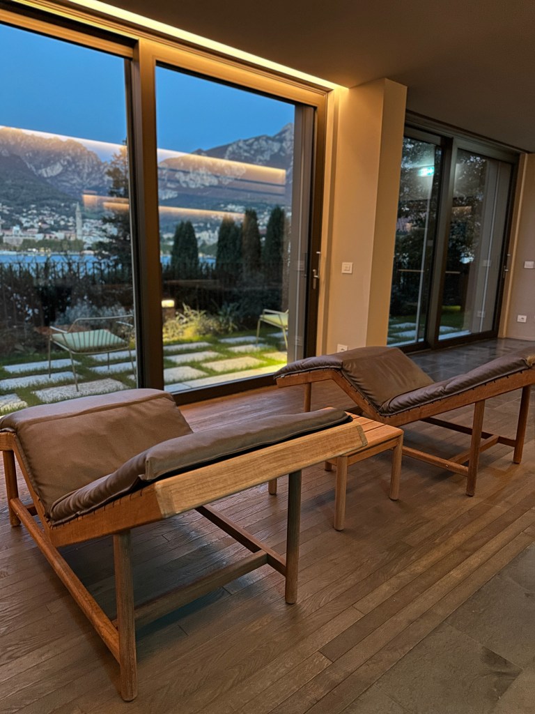 A cozy indoor setting featuring two wooden lounge chairs with cushions, positioned near large glass windows overlooking a scenic view of mountains and landscaped greenery.