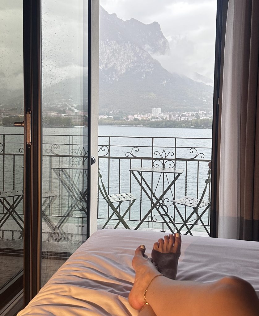A view from a hotel room showing a foot resting on a bed with a balcony overlooking a lake and mountains in the background, partially obscured by rain clouds.