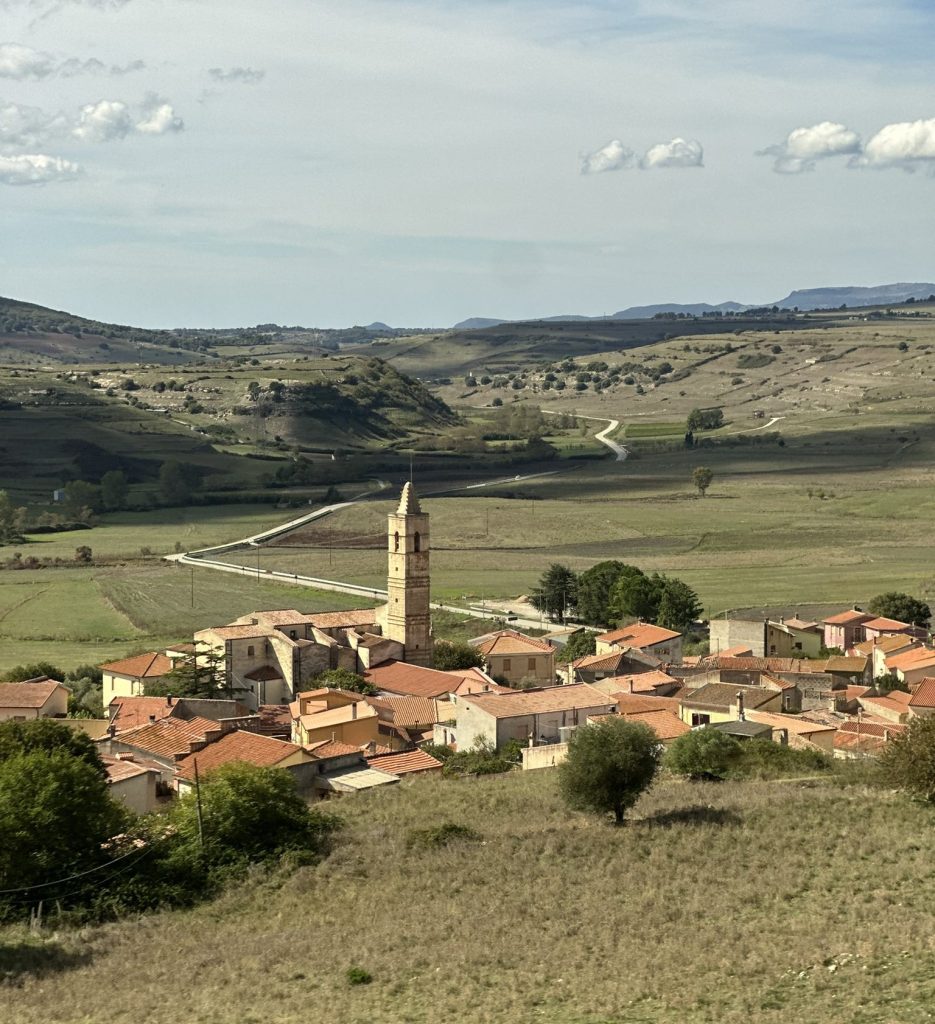 A picturesque village with red-roofed houses and a prominent bell tower, surrounded by green fields and rolling hills under a blue sky with scattered clouds.