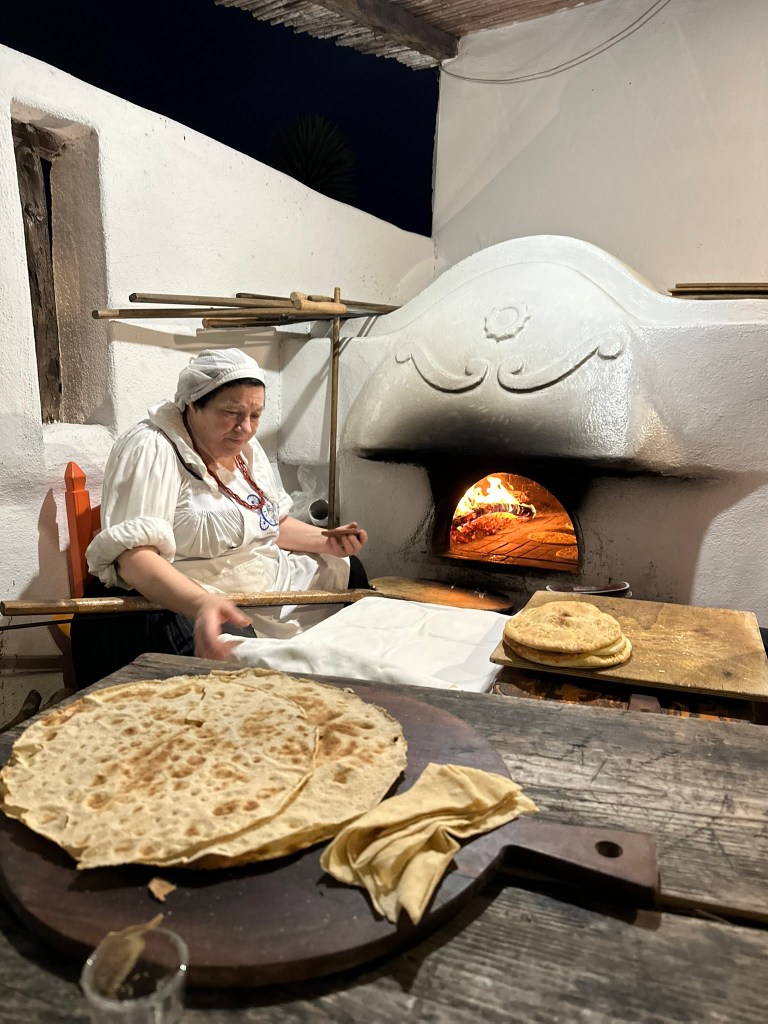 A woman in traditional attire prepares flatbreads in a rustic kitchen with a wood-fired oven, surrounded by freshly made flatbreads on a wooden table.