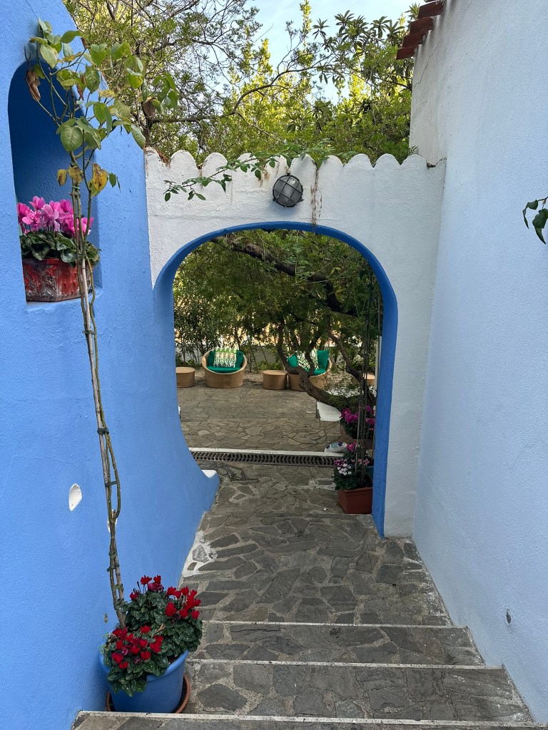 A scenic pathway leading through a blue archway, lined with flowering plants and stone steps, leading to a tranquil garden area.