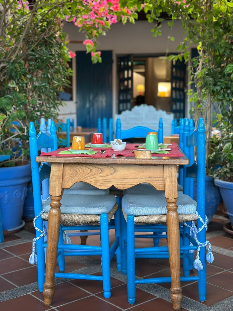 A wooden dining table surrounded by blue chairs, set with colorful cups and plates. The table is covered with a red tablecloth and is adorned with greenery and pink flowers in the background.