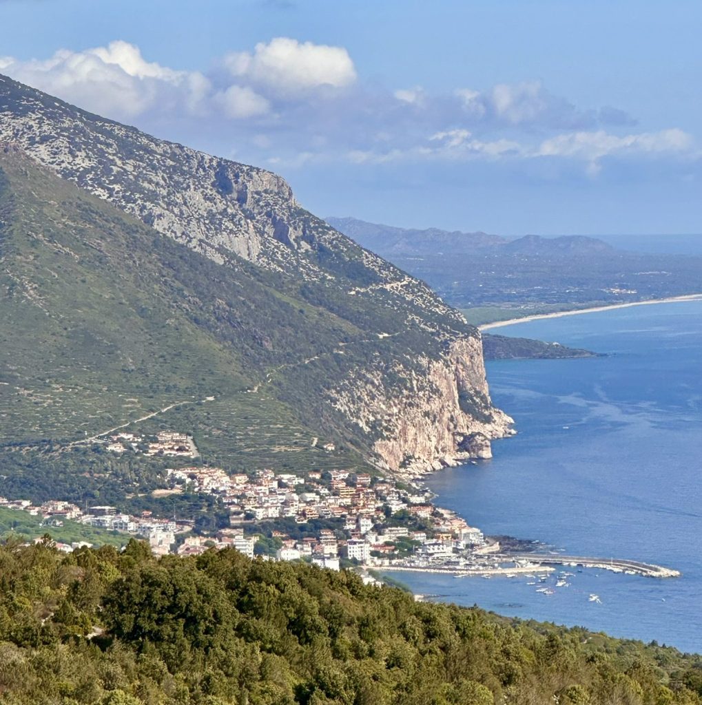 A coastal view featuring a small town nestled between green hills and a blue sea, with cliffs rising in the background and clouds in the sky.
