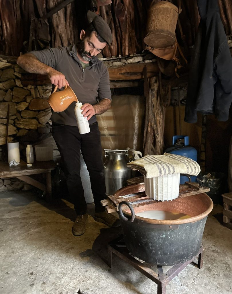 A man pouring liquid from a clay jug into a white container while standing next to a large copper basin in a rustic setting.