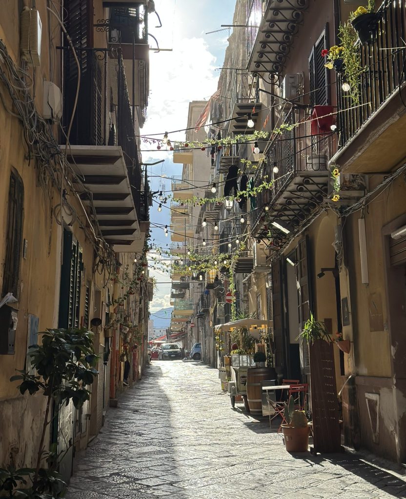 A narrow, sunlit street lined with buildings, adorned with hanging lights and greenery. A few potted plants are visible, and a small outdoor seating area is set up on the right side.