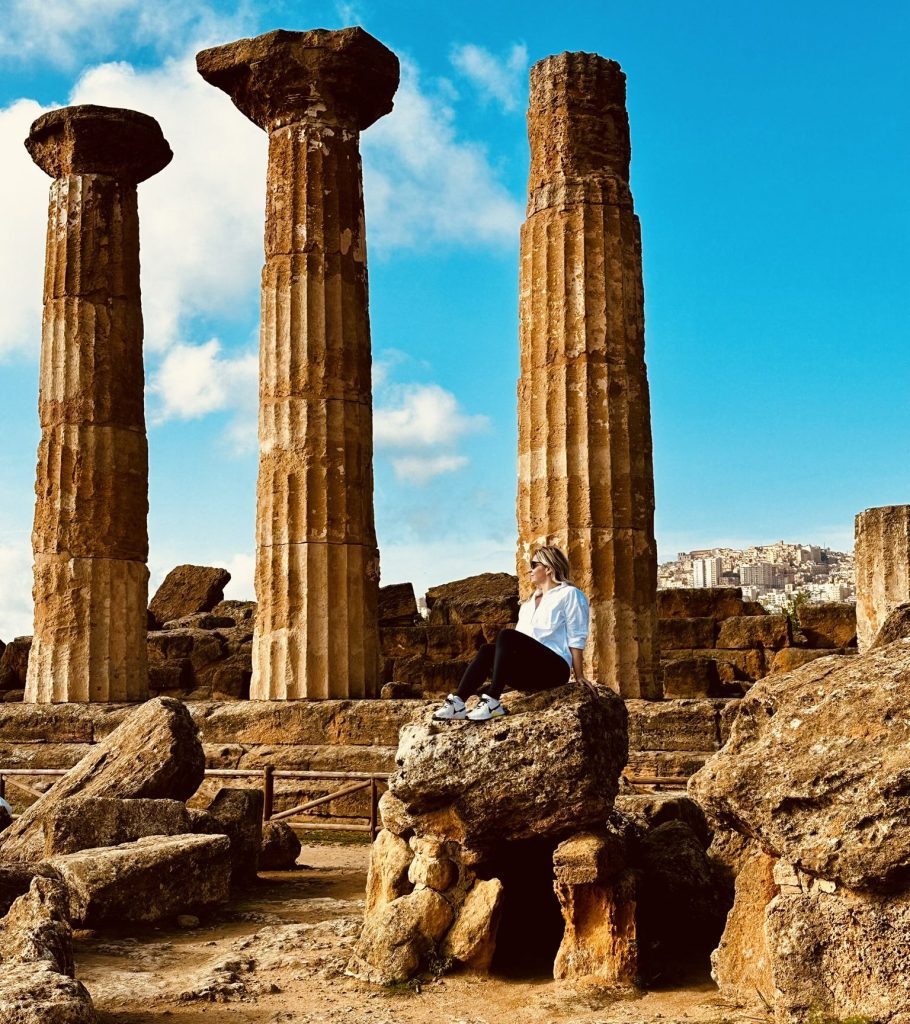 A woman sitting on a rock near ancient stone columns at a historical site, with a blue sky and distant buildings in the background.