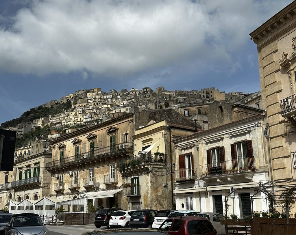 View of historic buildings in a town with a hillside covered in structures in the background under a cloudy sky.