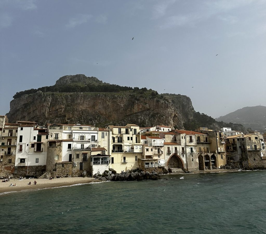 A coastal view featuring a rocky cliff towering over a quaint village with white buildings and red rooftops along the shoreline.