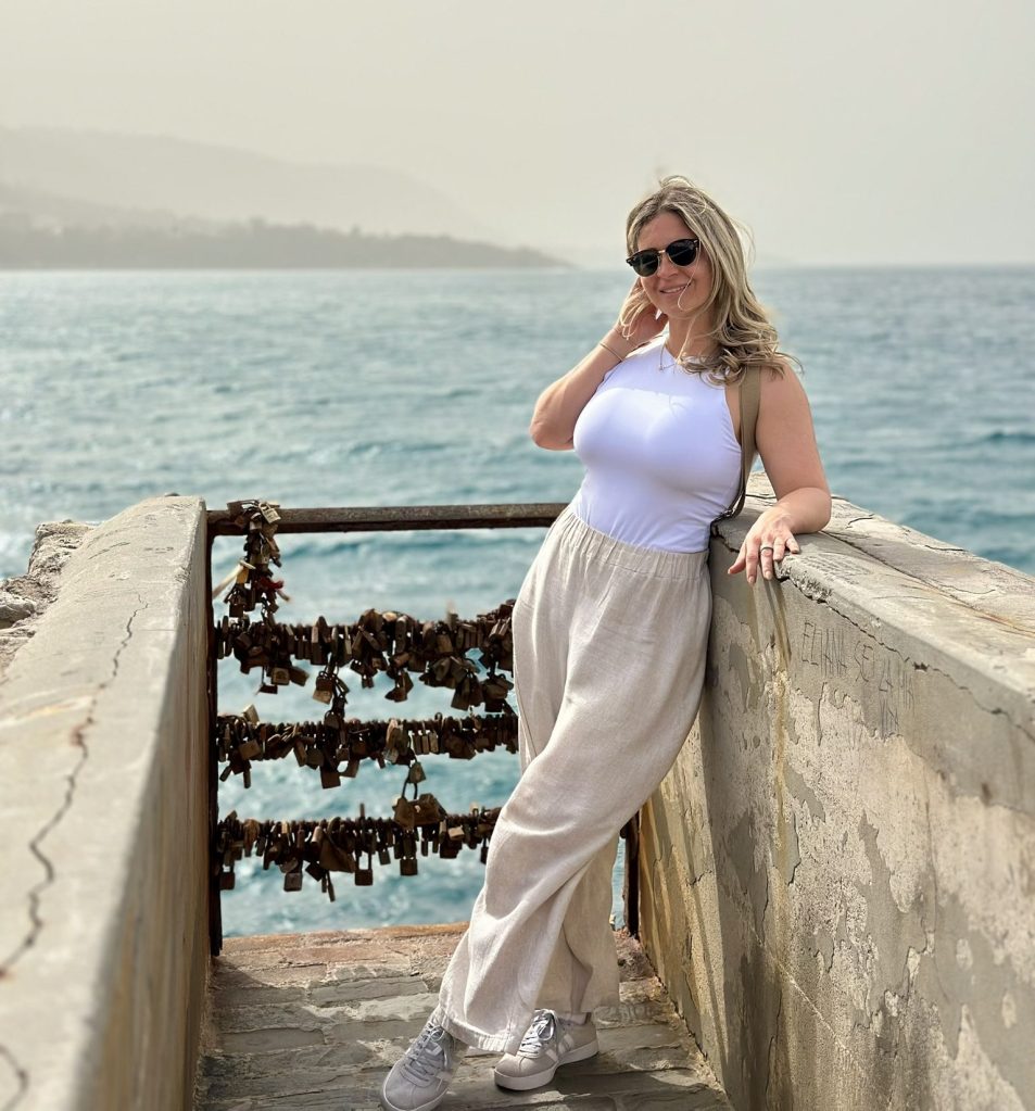 A woman poses casually on a railing near the water, wearing a white tank top and light-colored wide-leg pants, with sunglasses on. In the background, there is a view of the sea and a wall adorned with padlocks.