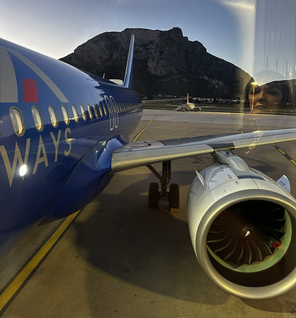 A view of an airplane on the tarmac with a mountain in the background, captured from a window. The image shows part of the airplane's wing and engine.
