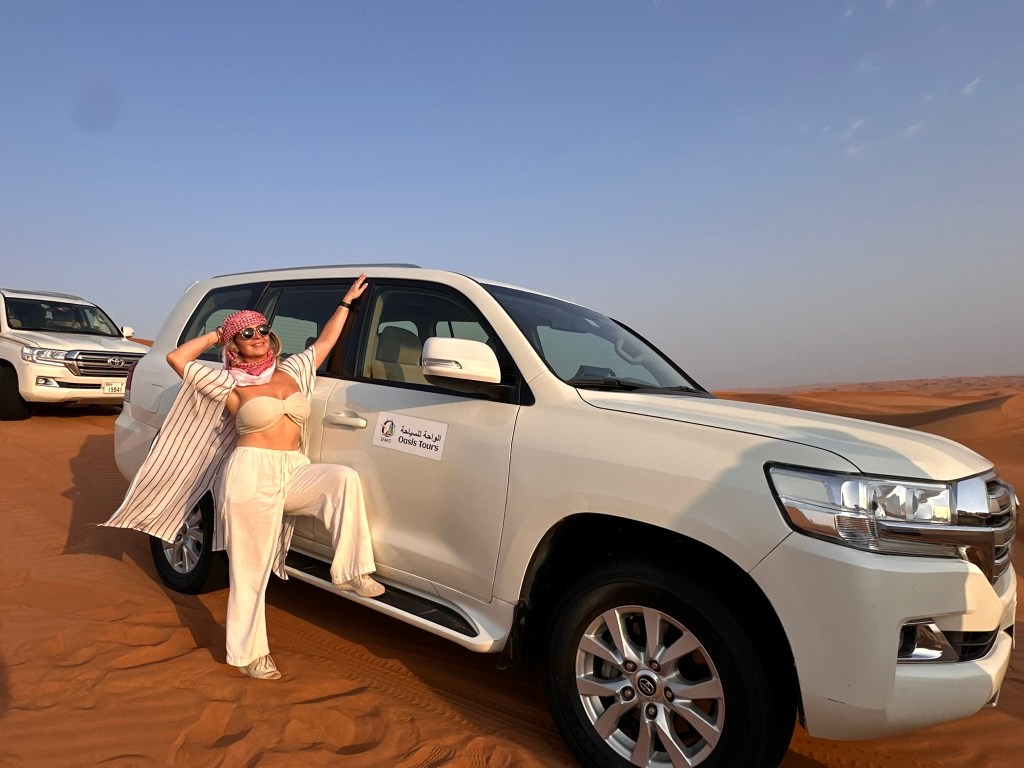 A person wearing a discreet outfit poses next to a white SUV in a desert landscape with sand dunes and a clear blue sky.