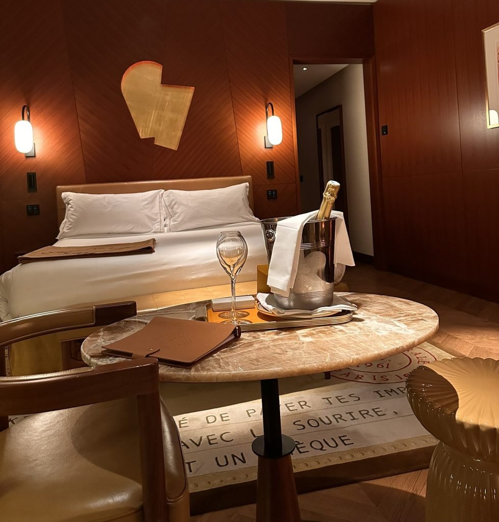 A luxurious hotel room featuring a king-sized bed with white linens, a marble table with a glass of champagne, an ice bucket, and decorative items, illuminated by stylish wall sconces.
