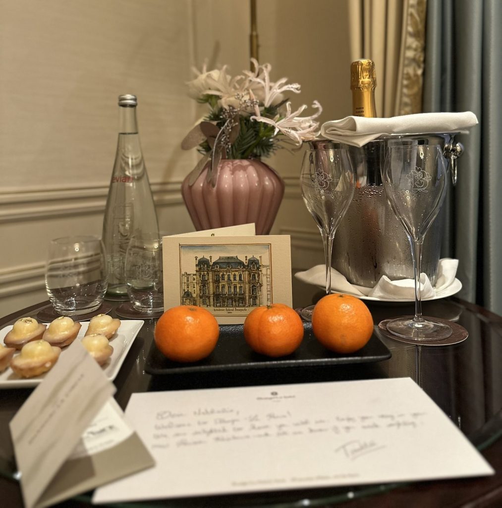 A table setting featuring a bottle of Evian water, champagne, two elegant glasses, fresh oranges, a plate of pastries, a greeting card with an illustration of a building, and a handwritten note, all arranged beside a pink vase with flowers.