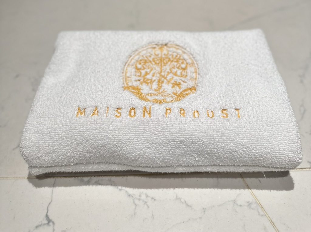 A folded white towel featuring the embroidered text 'MAISON PROUST' in gold.