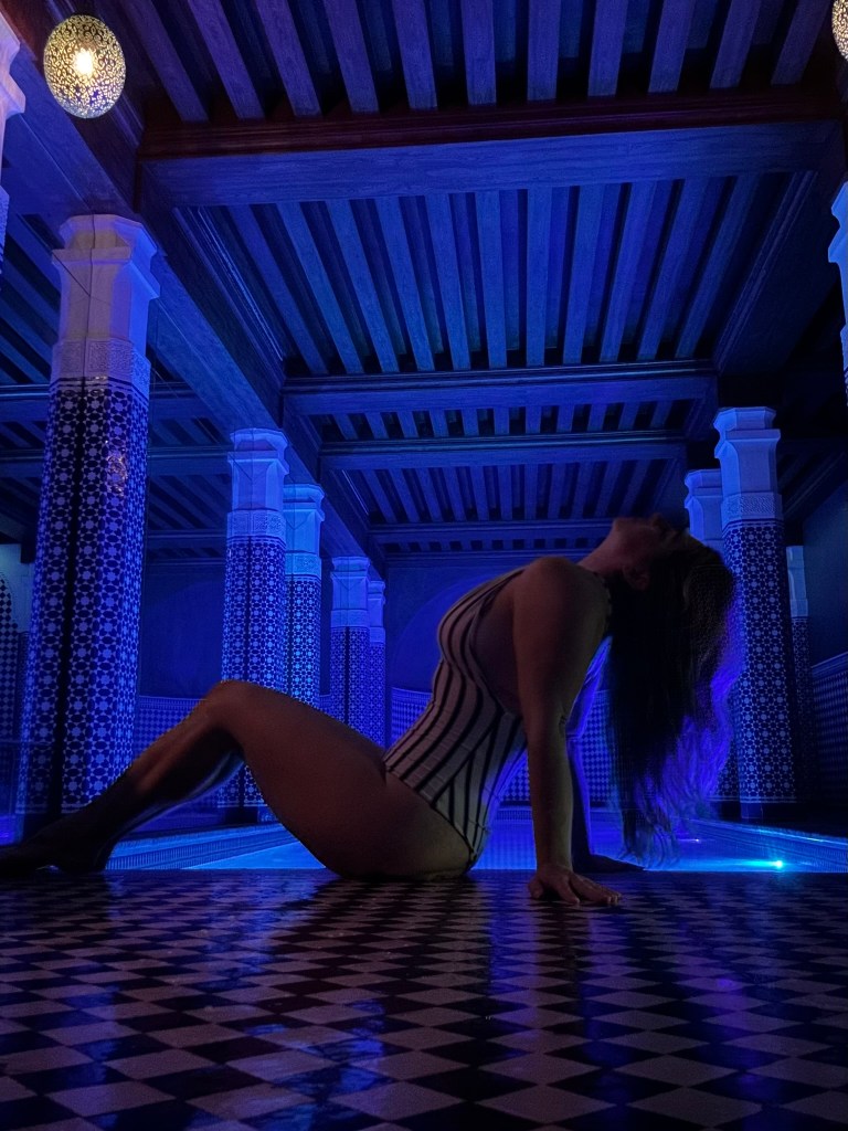 A woman in a striped swimsuit poses on the tiled floor of a dimly lit pool area with blue lights and ornate columns.
