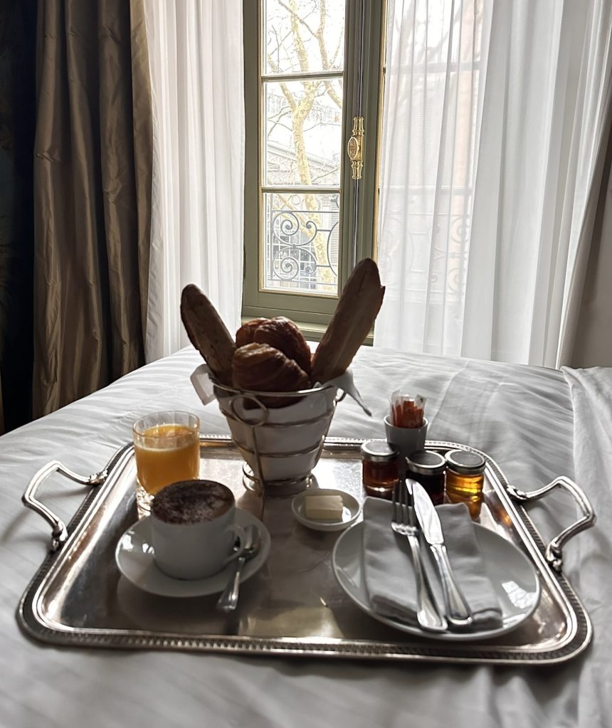 A silver tray with breakfast items including a cup of cappuccino, a glass of orange juice, a small bowl of butter, two jars of honey, pastries in a basket, and cutlery, set on a white bedspread with a window in the background.