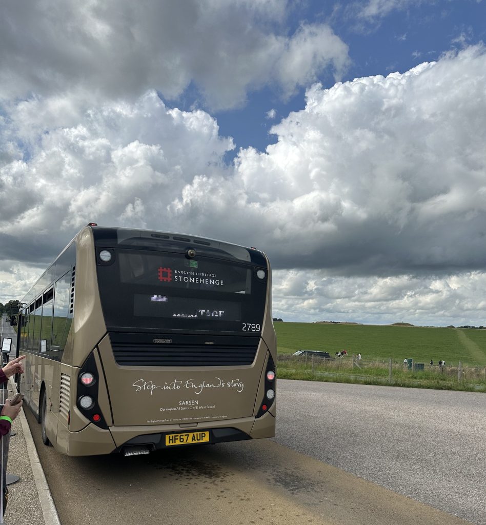 A beige bus with signage for Stonehenge parked on a rural road under a partly cloudy sky, with green fields in the background.